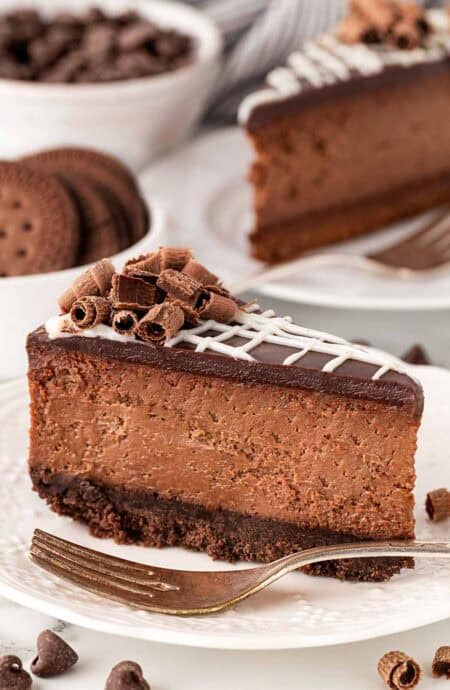 A slice of chocolate cheesecake with chocolate cookie crust topped with chocolate curls on a white plate with a fork next to it and cheesecake slices, chocolate, and cookies in the background