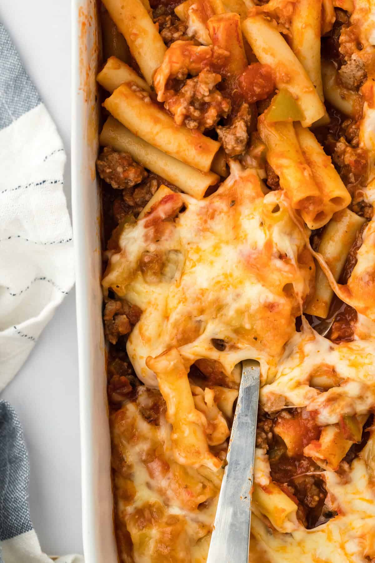 Spoon digging into baked ziti in a white baking dish