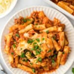 Easy baked ziti on a white plate sprinkled with parsley on white background with large dish of ziti and shredded cheese in background