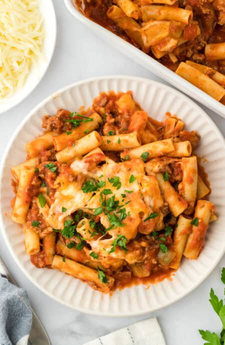 Easy baked ziti on a white plate sprinkled with parsley on white background with large dish of ziti and shredded cheese in background