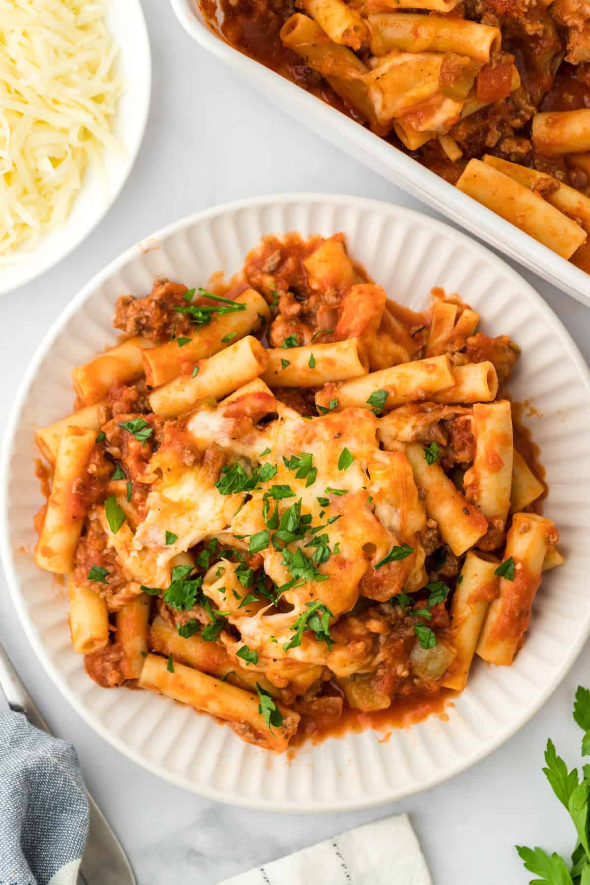 Easy baked ziti on a white plate sprinkled with parsley on white background with large dish of ziti and shredded cheese in background