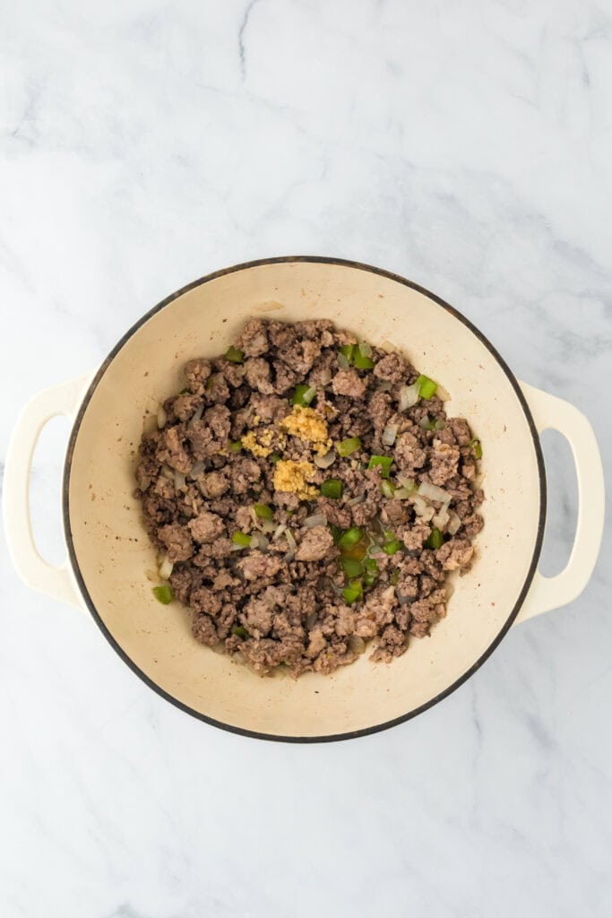 Browned meat in a large pot with garlic added as well on white background.