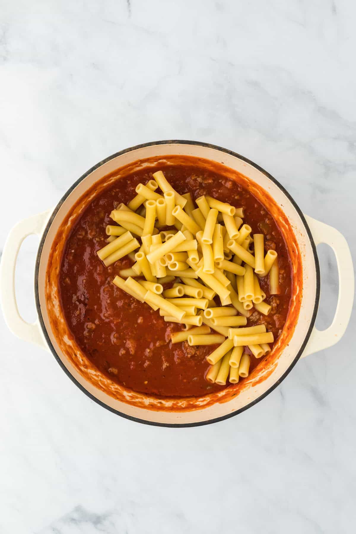 Cooked ziti pasta added to a tomato sauce in a white pot on white background