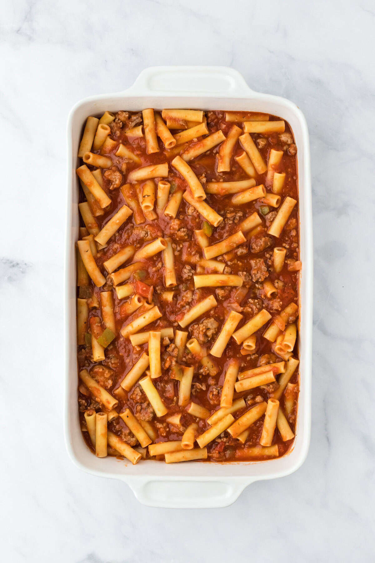 Baked ziti filling added to a white baking dish on a white background before baking