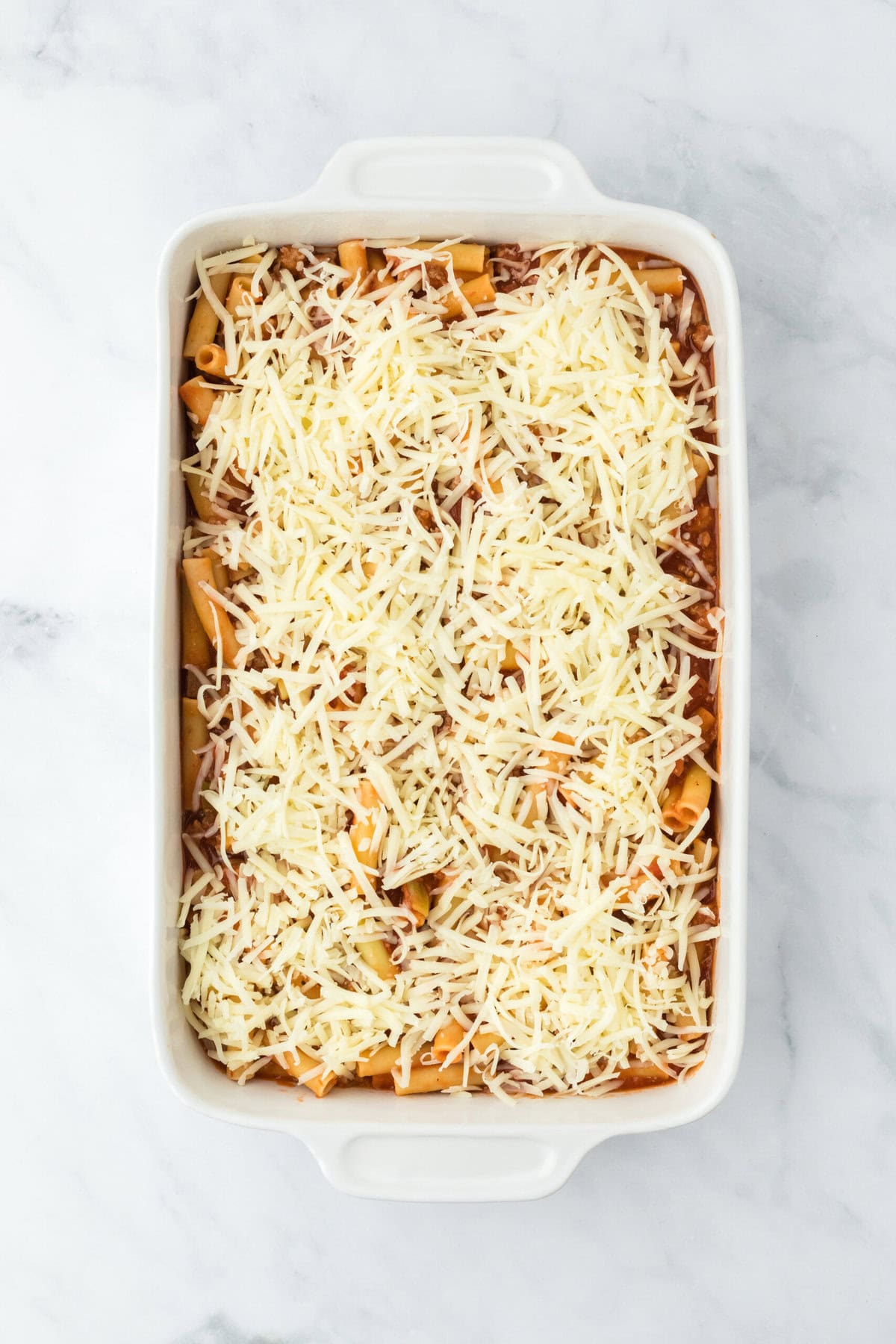 Shredded mozzarella cheese added on top of baked ziti before baking on white background