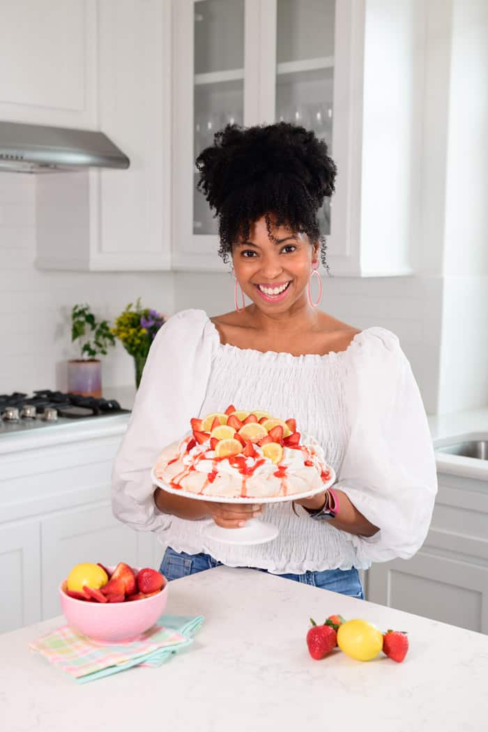 Jocelyn Delk Adams holding a strawberry lemon pavlova in a white kitchen and smiling