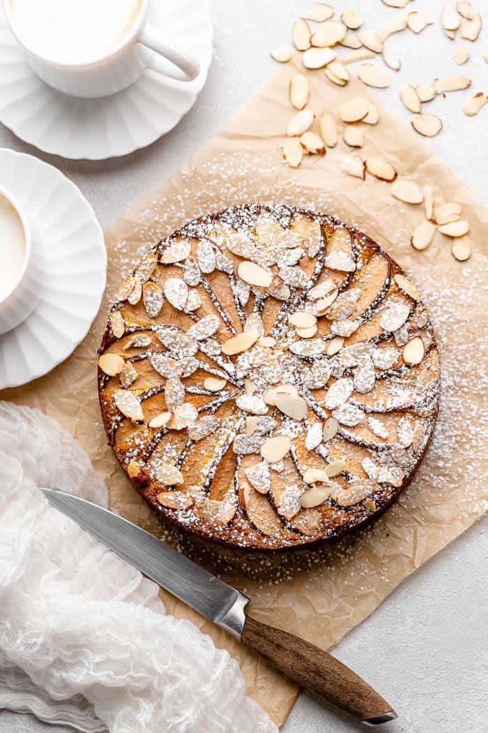 A whole one layer polenta cake with almonds on top against parchment paper with cups of tea
