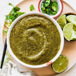 A large white bowl of salsa verde with limes, cilantro and peppers around it
