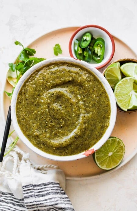 A large white bowl of salsa verde with limes, cilantro and peppers around it
