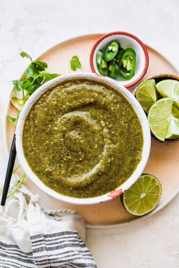 A large white bowl of salsa verde with limes, cilantro and peppers around it