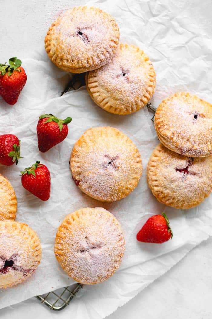 Strawberry hand pies scattered on white parchment paper with strawberries scattered about