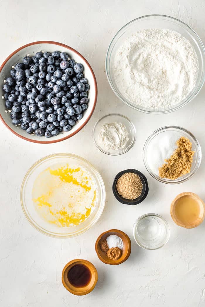 Delicious ingredients like fresh blueberries, sugars, spices, cake mix, melted butter and flour in bowls against white background