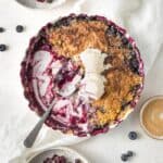 A casserole dish filled with blueberry cobbler being scooped out with a large spoon and ice cream on top