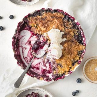 A casserole dish filled with easy blueberry cobbler recipe being scooped out with a large spoon and ice cream on top against a white background