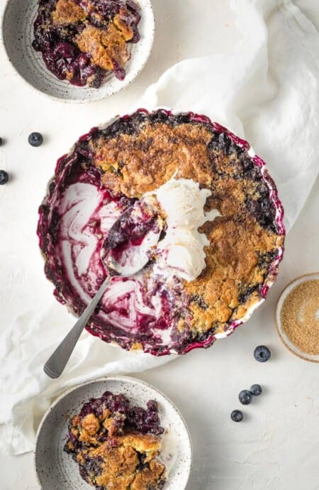 A casserole dish filled with easy blueberry cobbler recipe being scooped out with a large spoon and ice cream on top against a white background