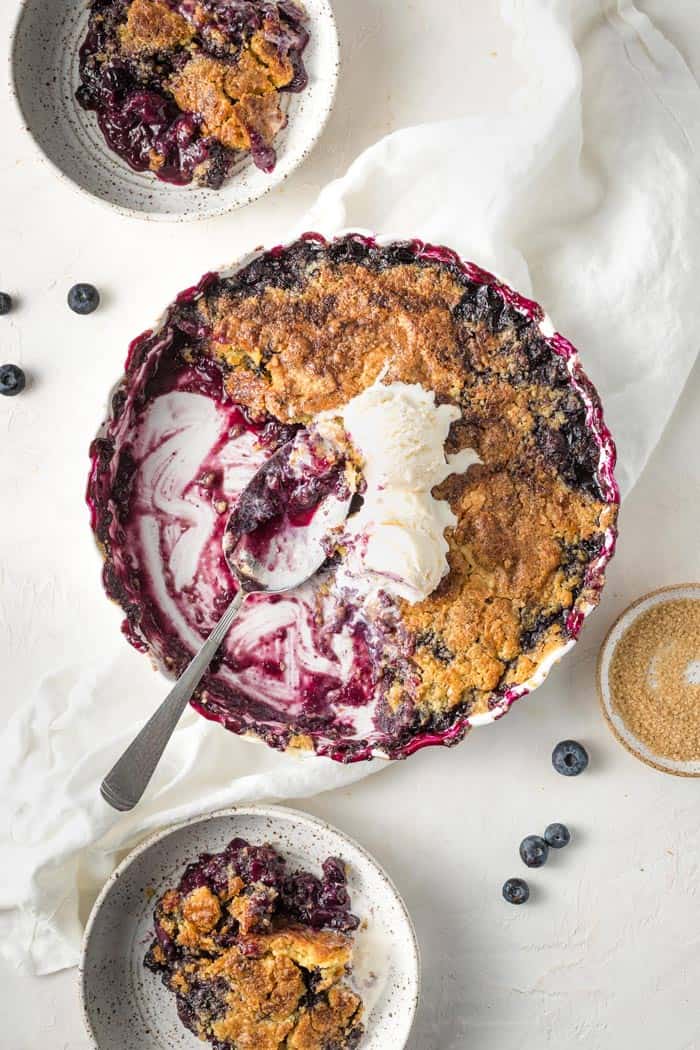 A casserole dish filled with blueberry cobbler being scooped out with a large spoon and ice cream on top