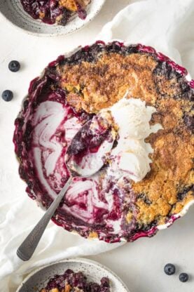 A close up of blueberry cobbler scooped out of a round pan with ice cream on top