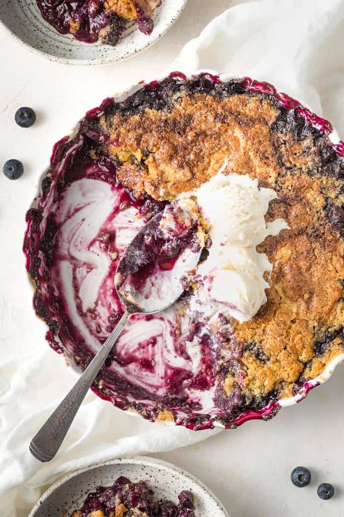 A close up of simple blueberry cobbler scooped out of a round pan with ice cream on top against a white background