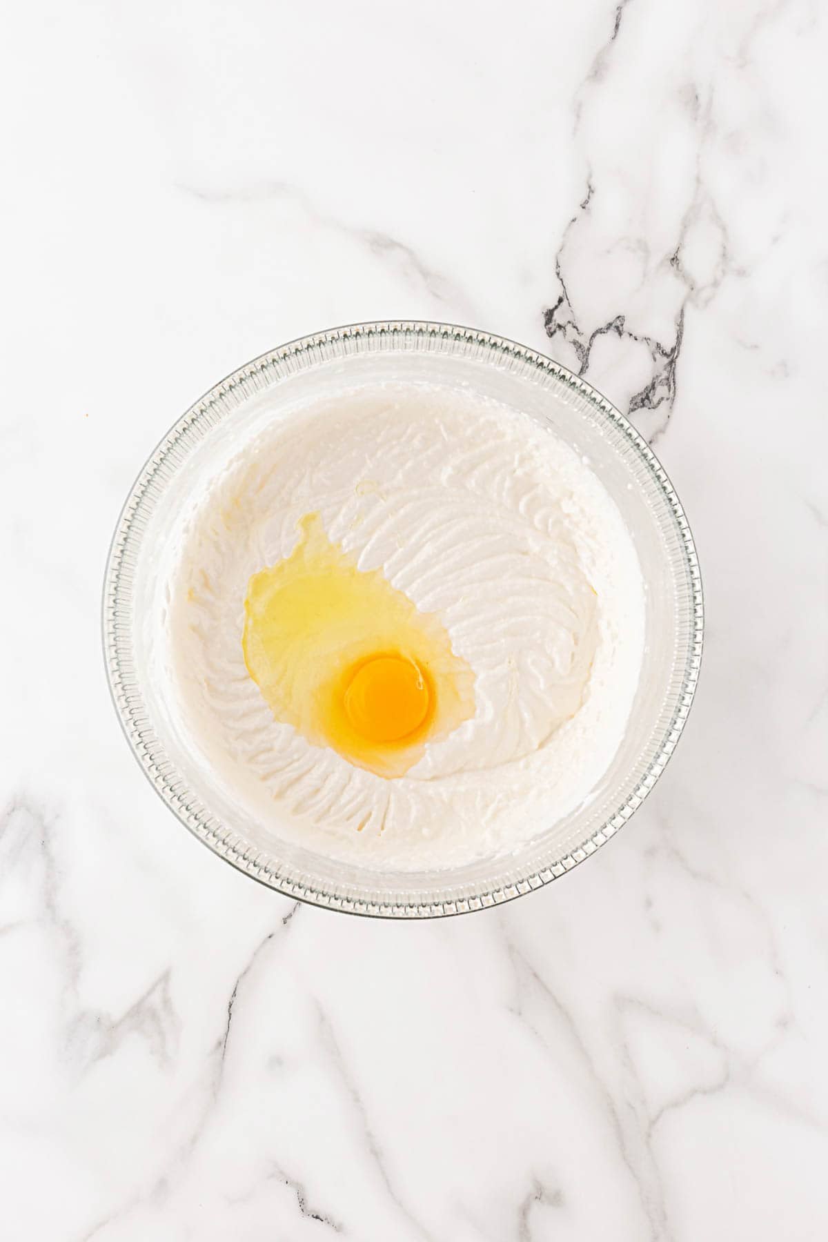 A mixing bowl with whipped batter and an egg cracked into the center, ready to be mixed
