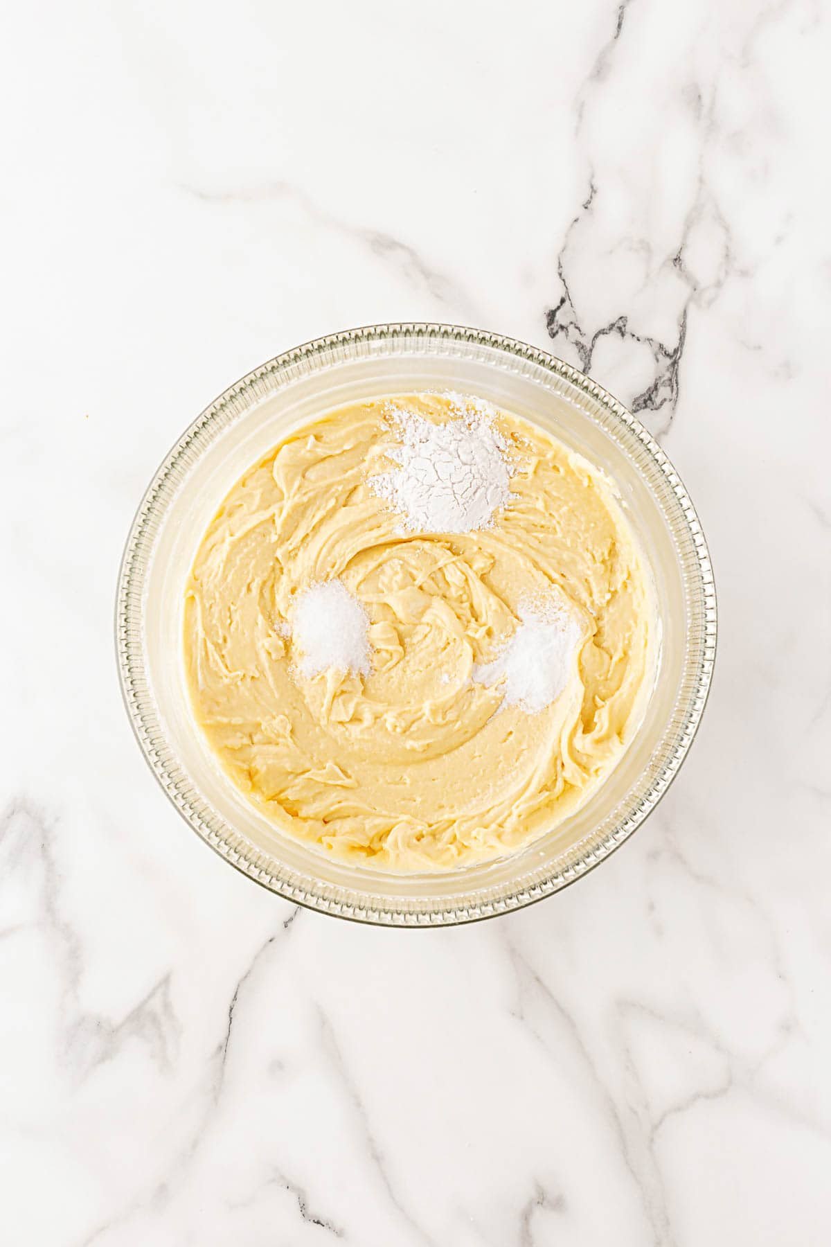 A glass mixing bowl filled with smooth yellow batter topped with baking powder, salt and baking soda