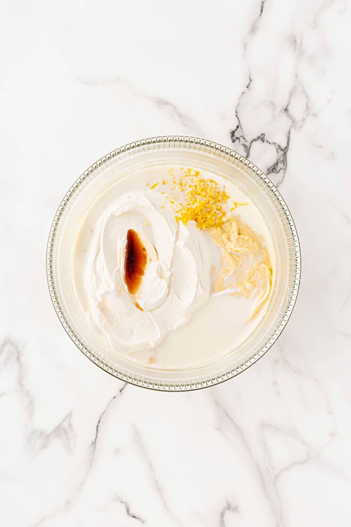 A glass mixing bowl filled with a smooth white batter with sour cream, milk, lemon zest, lemon and vanilla extracts added
