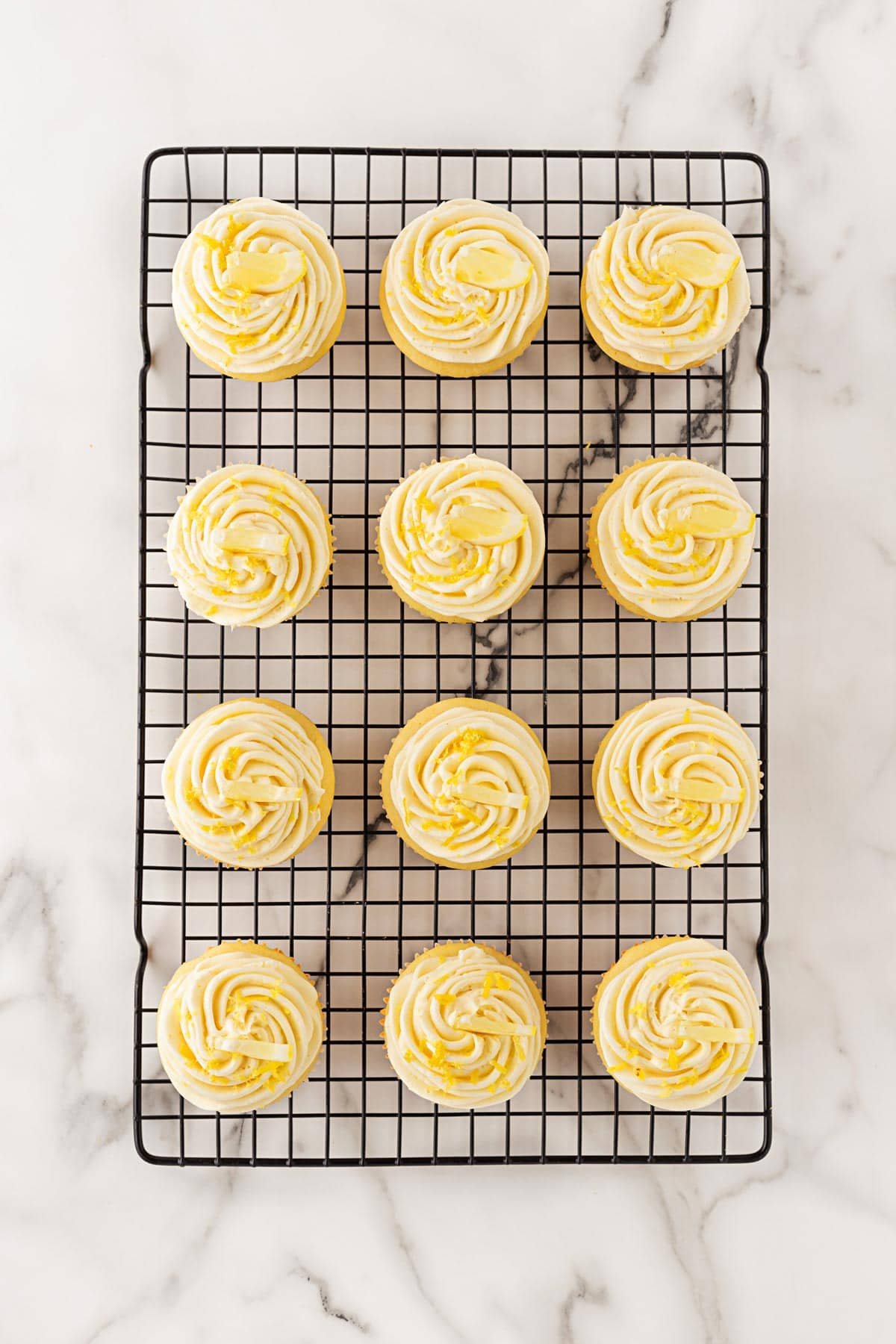 Frosted lemon cupcakes cooling on a black wire rack, swirled with lemon buttercream and zest