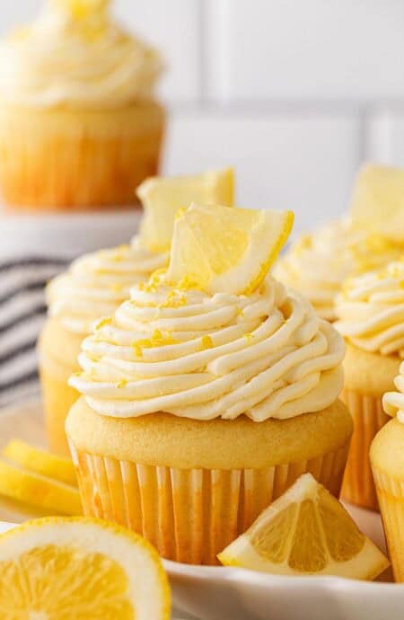 A plate full of frosted lemon cupcakes decorated with lemon wedges and zest