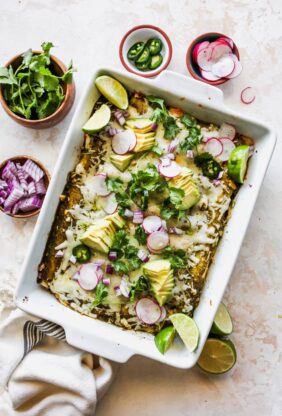 A casserole dish filled with chicken enchiladas topped with red onion, limes and cilantro