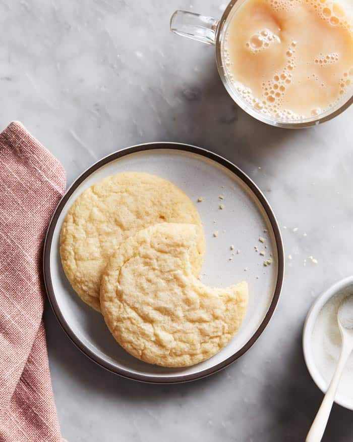 Two lemon tea cake cookies on a white plate with a bite taken along with some coffee