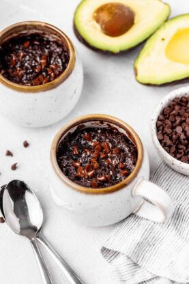 Two brownie in a mug servings ready to enjoy with avocado in the background