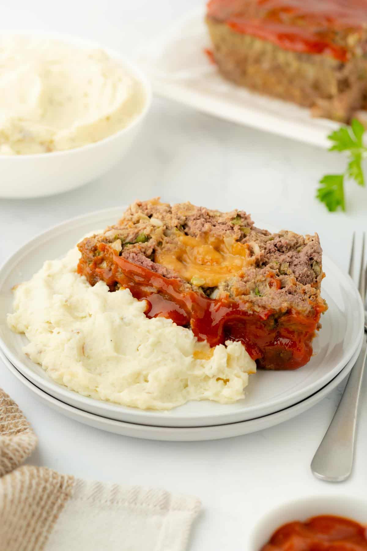 A serving plate with mashed potatoes and a slice of stuffed meatloaf, with more mash and meatloaf in the background