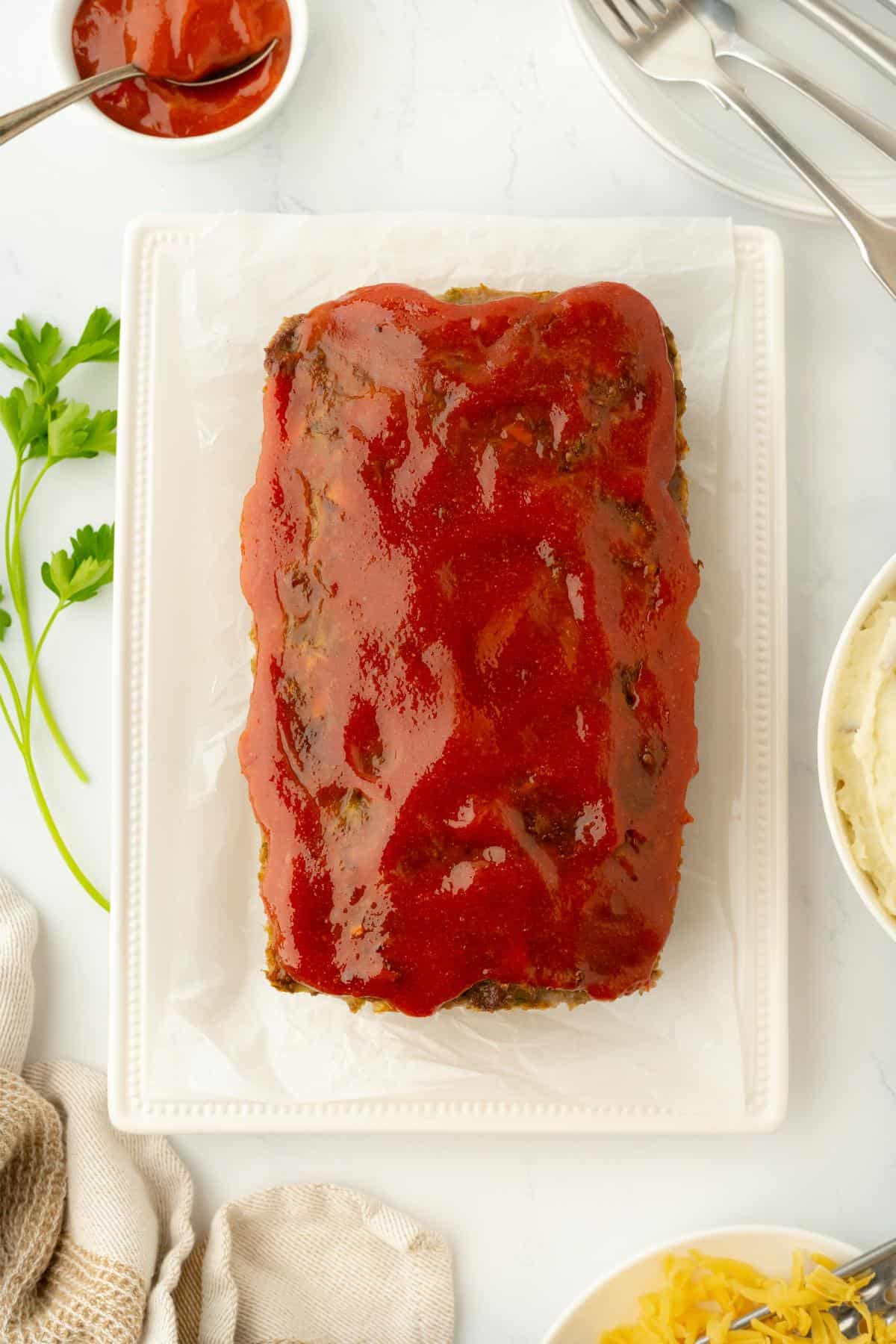 A fully baked glazed stuffed meatloaf served on a rectangular plate with parsley garnish, mashed potatoes, and shredded cheese on the side