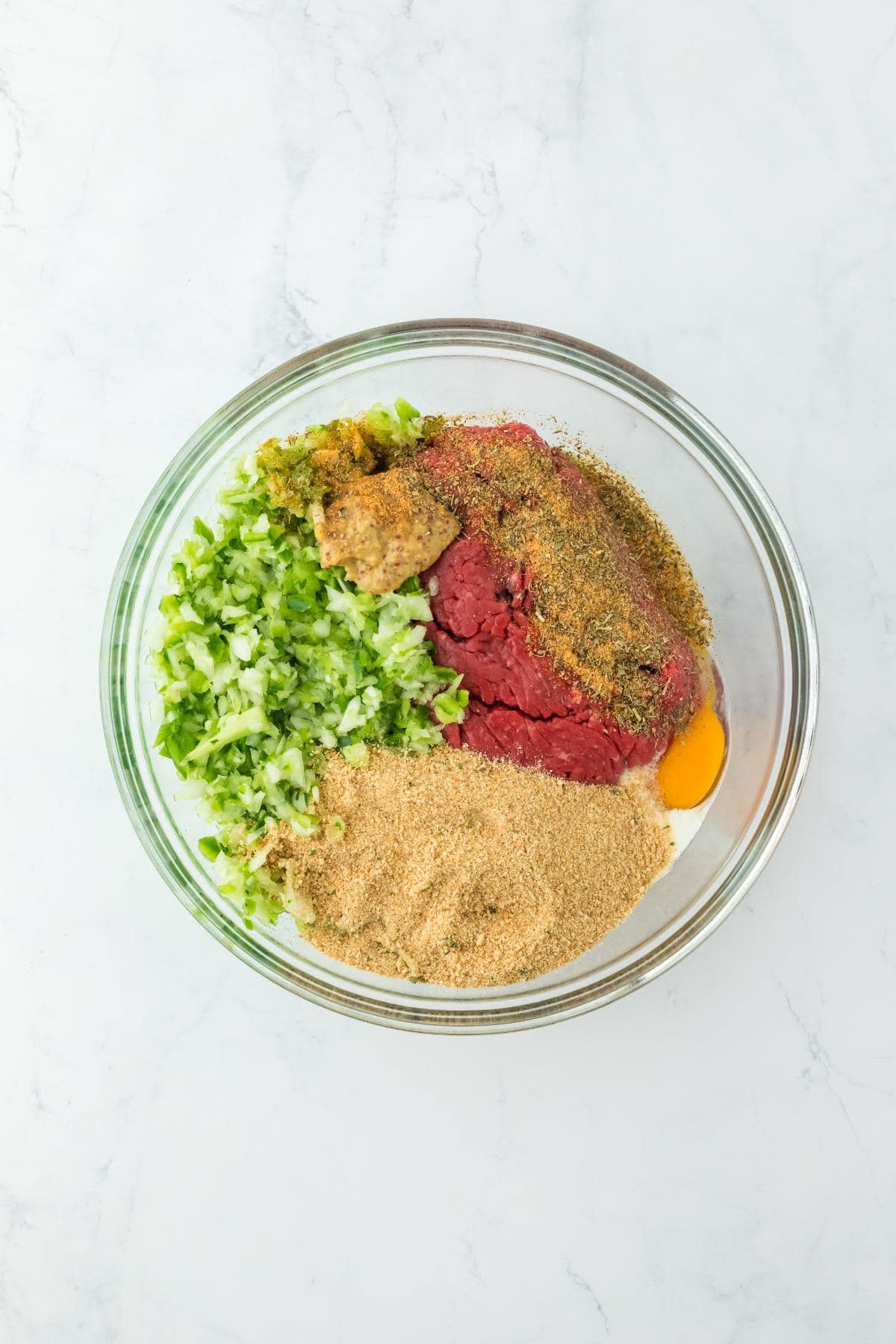 A large glass bowl with ground beef, chopped vegetables, breadcrumbs, mustard, egg, and seasoning