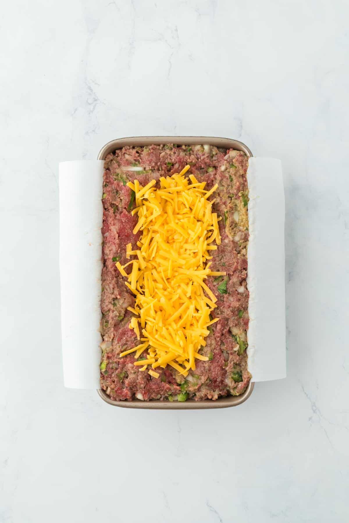 A loaf pan with meatloaf mixture, showing a layer of shredded cheese in the center