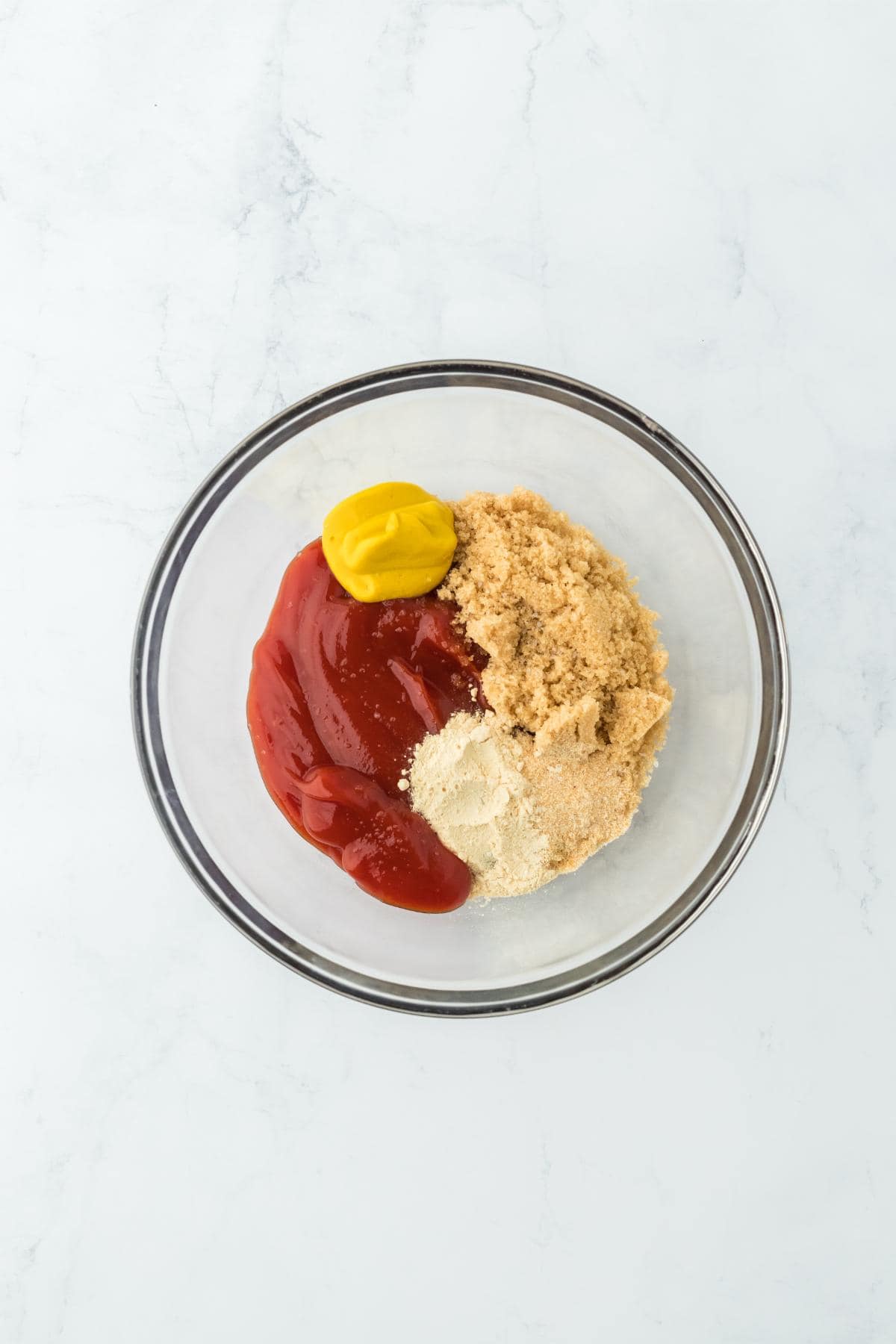 A glass bowl containing ketchup, mustard, brown sugar, and spices before mixing