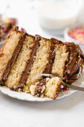 A close up of a slice of yellow cake with chocolate frosting on a white plate with a fork going through it