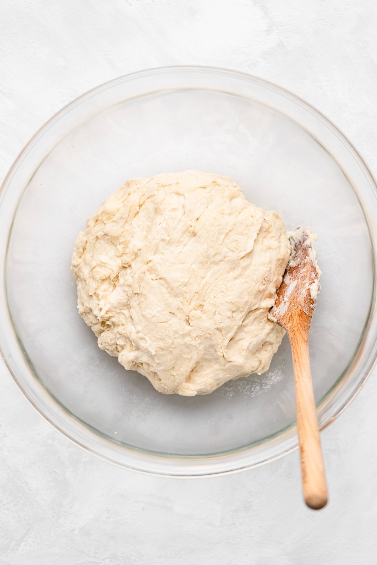 A ball of bread dough resting in a large glass bowl, with a wooden spoon partially covered in dough