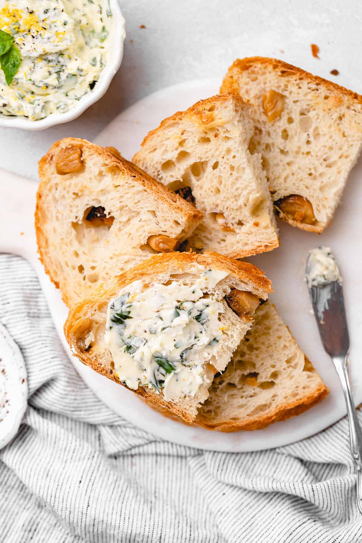 Multiple slices of roasted garlic no knead bread on a marble board, with one slice generously spread with basil butter