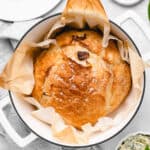 A golden-brown round loaf of bread baked in a Dutch oven, topped with flaky sea salt and parchment lining