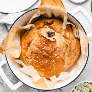 A golden-brown round loaf of bread baked in a Dutch oven, topped with flaky sea salt and parchment lining