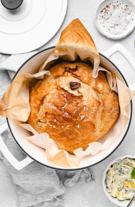 A golden-brown round loaf of bread baked in a Dutch oven, topped with flaky sea salt and parchment lining