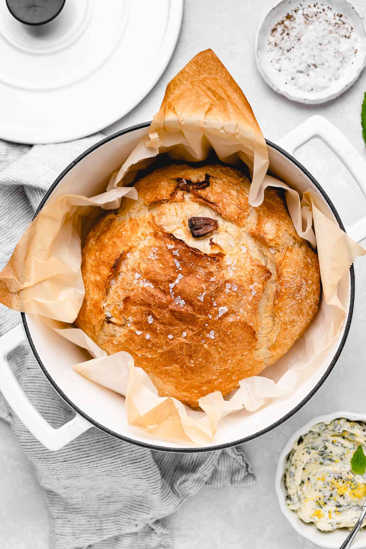 A golden-brown round loaf of bread baked in a Dutch oven, topped with flaky sea salt and parchment lining