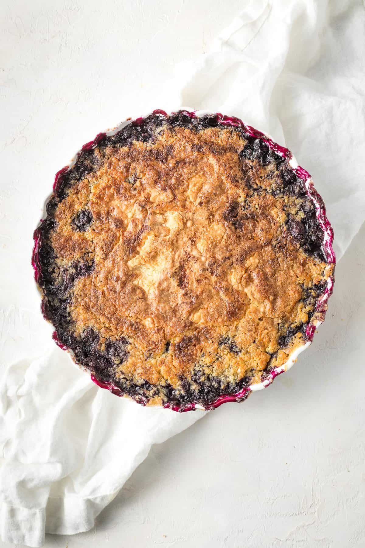 An easy blueberry cobbler in a round baking dish after baked until golden on a white background