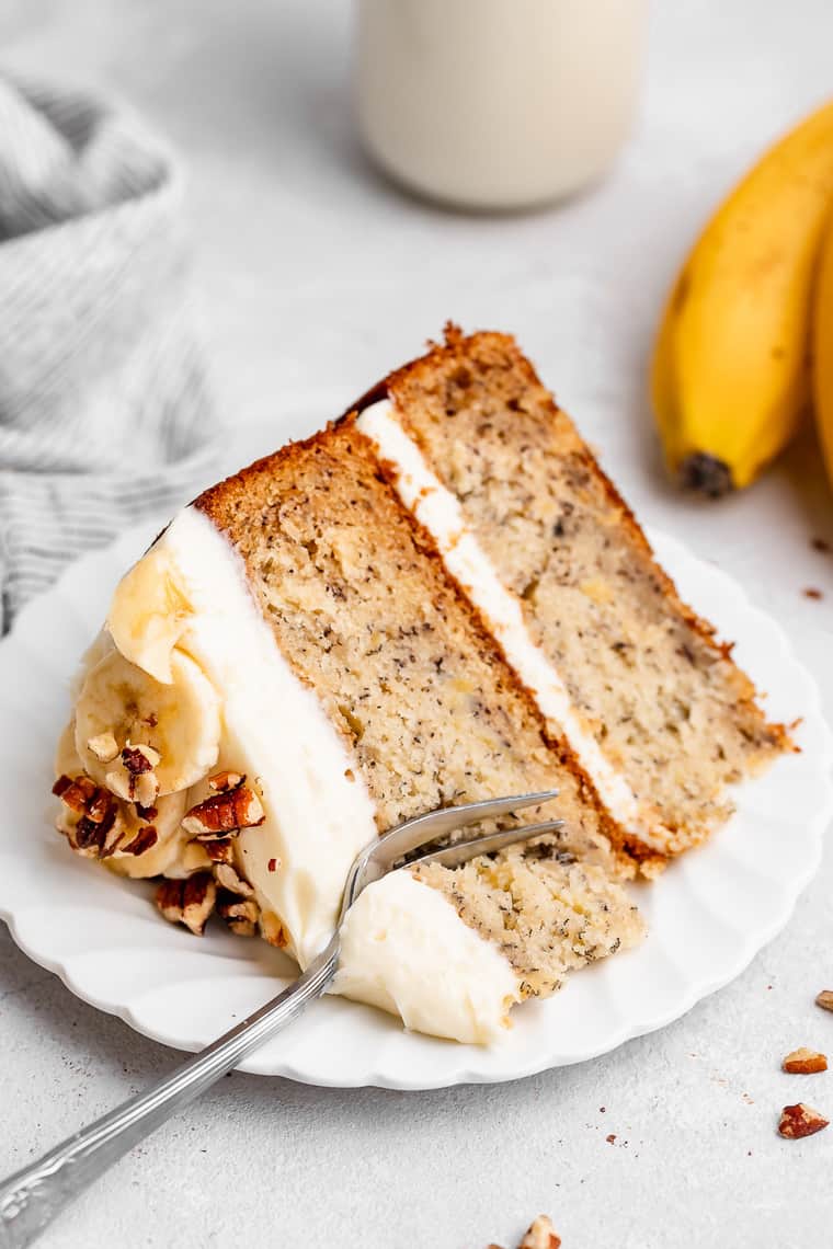 A large slice of banana two layer cake on a white plate with a fork slicing through it
