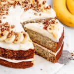 A slice of banana cake with cream cheese frosting being pulled out of whole cake with bananas in the background