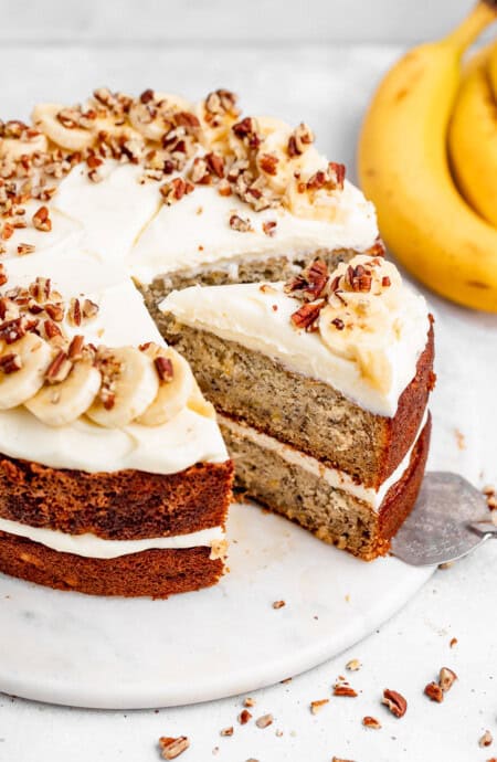 A slice of banana cake with cream cheese frosting being pulled out of whole cake with bananas in the background