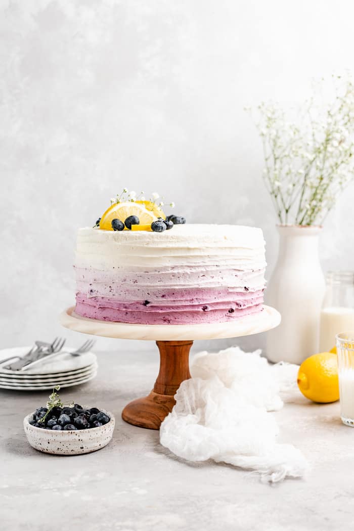 A layer cake filled with lemon flavor and blueberries topped with fresh fruit on a wooden cake stand