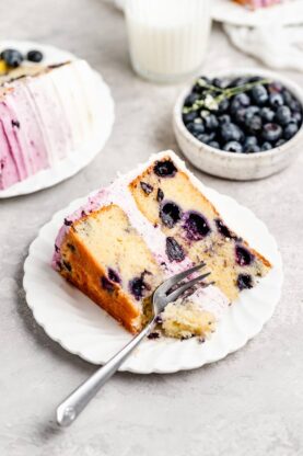 A slice of lemon layer cake with blueberries on a white plaate with a fork eating