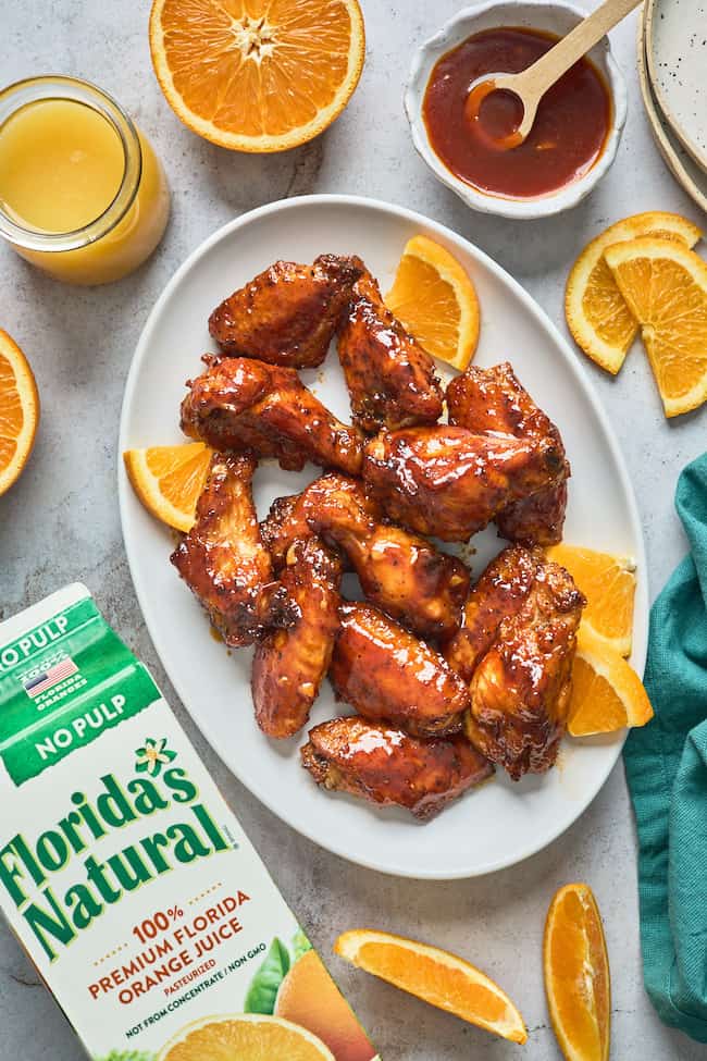 A white platter filled with honey bbq wings with orange slices against a gray background with orange wedges and orange juice carton