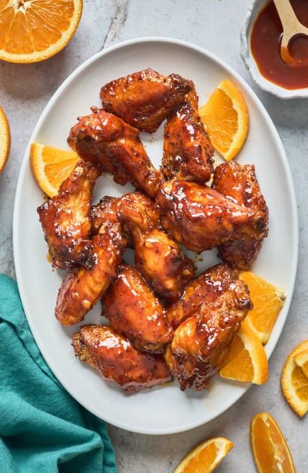 A delicious white platter filled with honey bbq wings with orange slices and honey in a container nearby